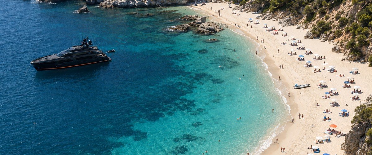 Aerial view of a curved sandy beach and clear water below rocky hills, a dark yacht anchored offshore - illustrative context