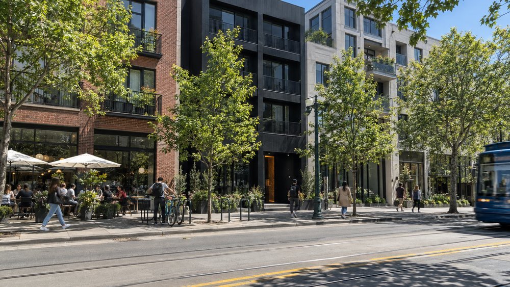 People walking and sitting at cafe tables along a tree-lined street, a black building between brick and stone neighbours - illustrative context