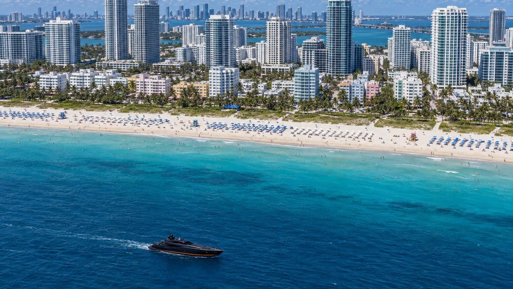 Aerial view of a coastal city with high-rises behind a busy beach, a dark boat crossing the water - illustrative context